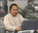 A man wears a headset with microphone while sitting at a desk using a laptop computer. 