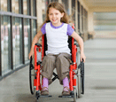 A young, smiling girl using a wheelchair, rolling down a walkway.