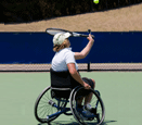 A person playing tennis while sitting in a wheelchair that has wheels angled in at the top.