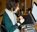 A woman sitting at a desk using a laptop computer and wearing a headset with microphone and some sort of wrist attachments.
