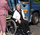 An older woman with grey hair sitting in a wheelchair that is on a lift attached to a van.