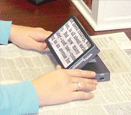 a rectangular device sits on top of a newspaper and magnifies the text of the newspaper. A person is sitting and looking at the device. 