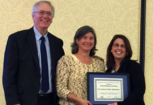 Three people stand together, two women and one man. They are wearing business type clothes. The women hold a framed certificate.  